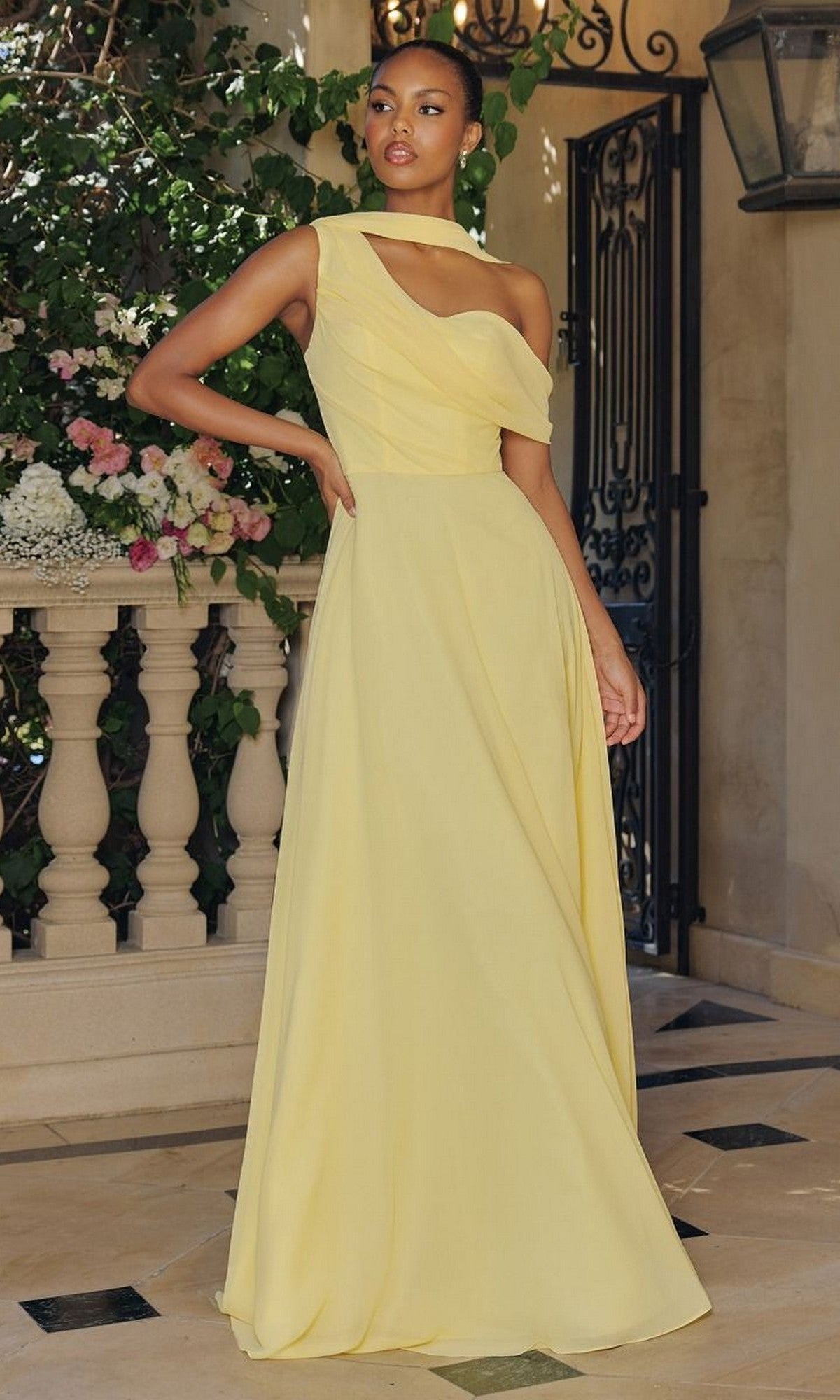 A woman stands on a stone patio in the Amoris T716 Asymmetric Long A-Line Prom Dress, its light yellow hue and one-shoulder design highlighted by flowers and greenery that enhance the elegance of this stunning evening gown.